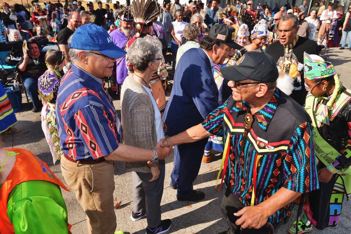Two men in patterned shirts and baseball caps shake hands amidst a crowd at an outdoor American Indian heritage event.