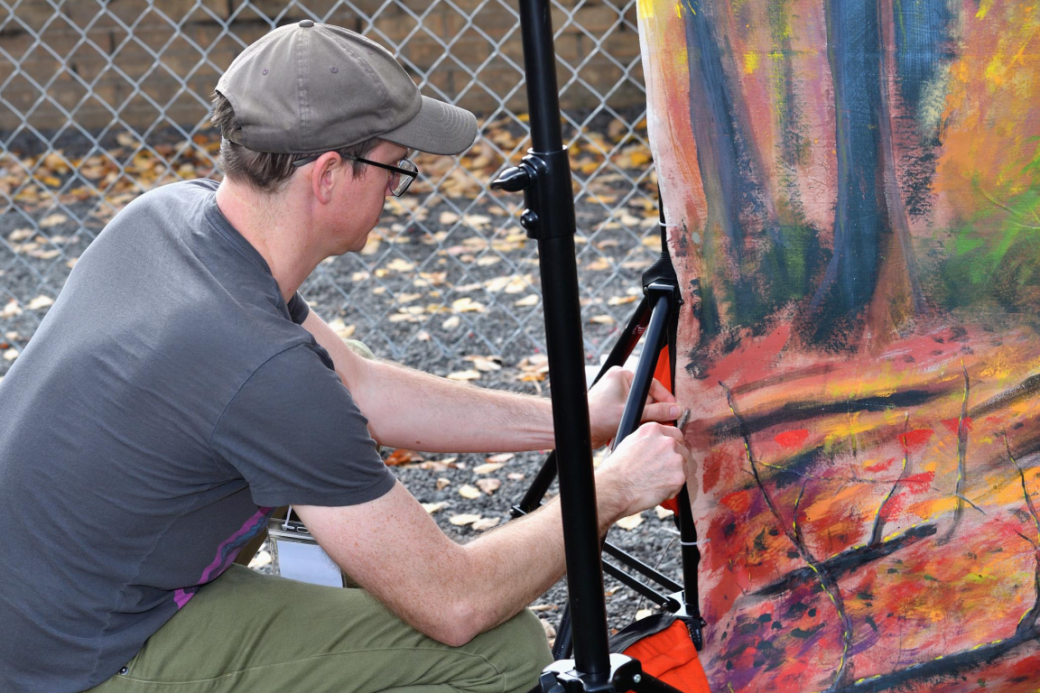 A man in a baseball cap and glasses adjusts the back of a large, colorful autumn-themed display panel outdoors.