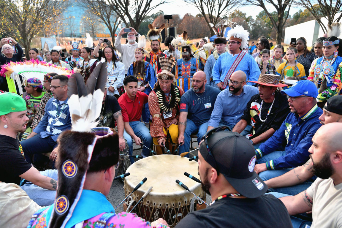 A large group of people, many in traditional American Indian regalia and headdresses, gather around a large drum and sing during an outdoor event.