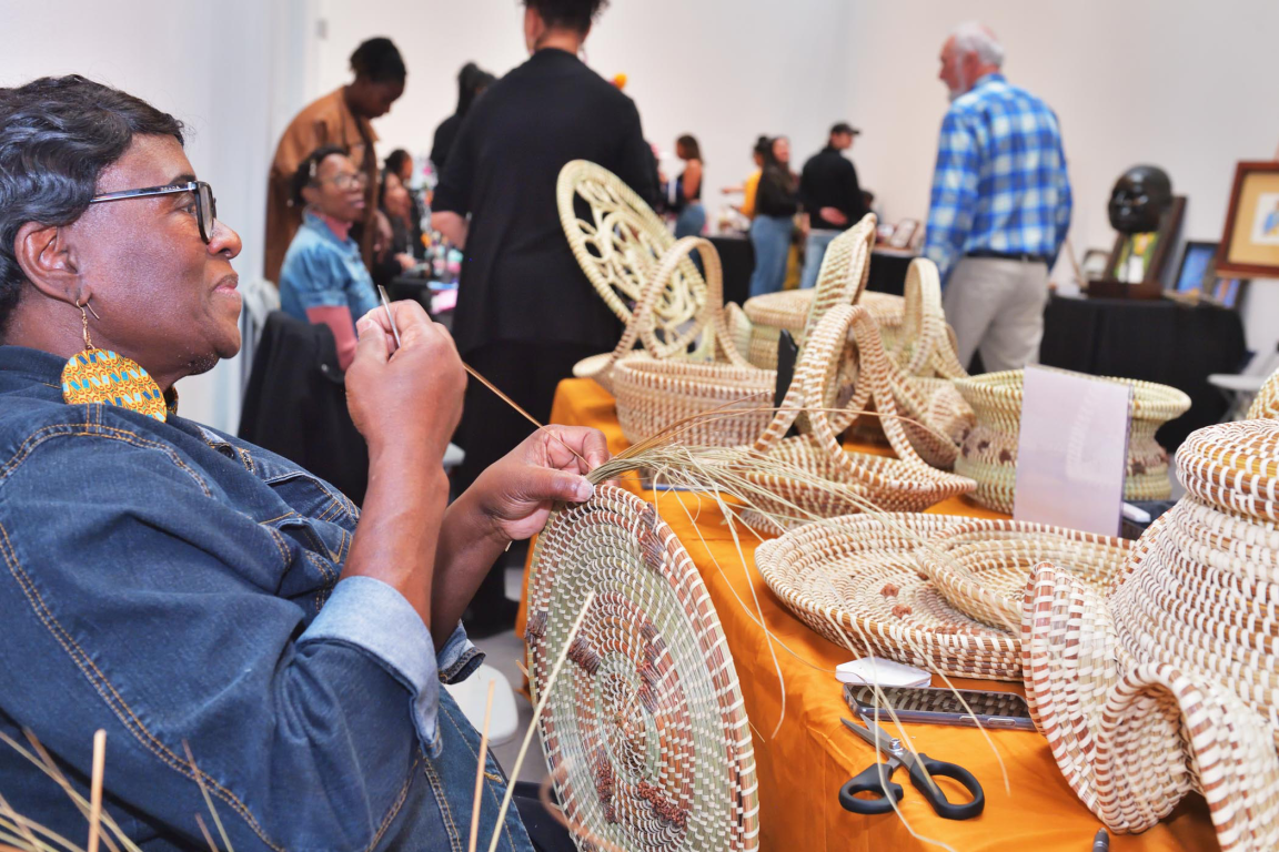 A woman in a denim jacket weaves a sweetgrass basket at a table displaying finished woven baskets of various shapes and sizes while museum visitors mingle in the background.