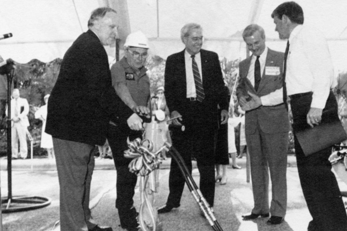 Black-and-white photograph of several men in suits and a construction worker gathered under a tent, participating in a ceremonial groundbreaking using a drill adorned with a ribbon.