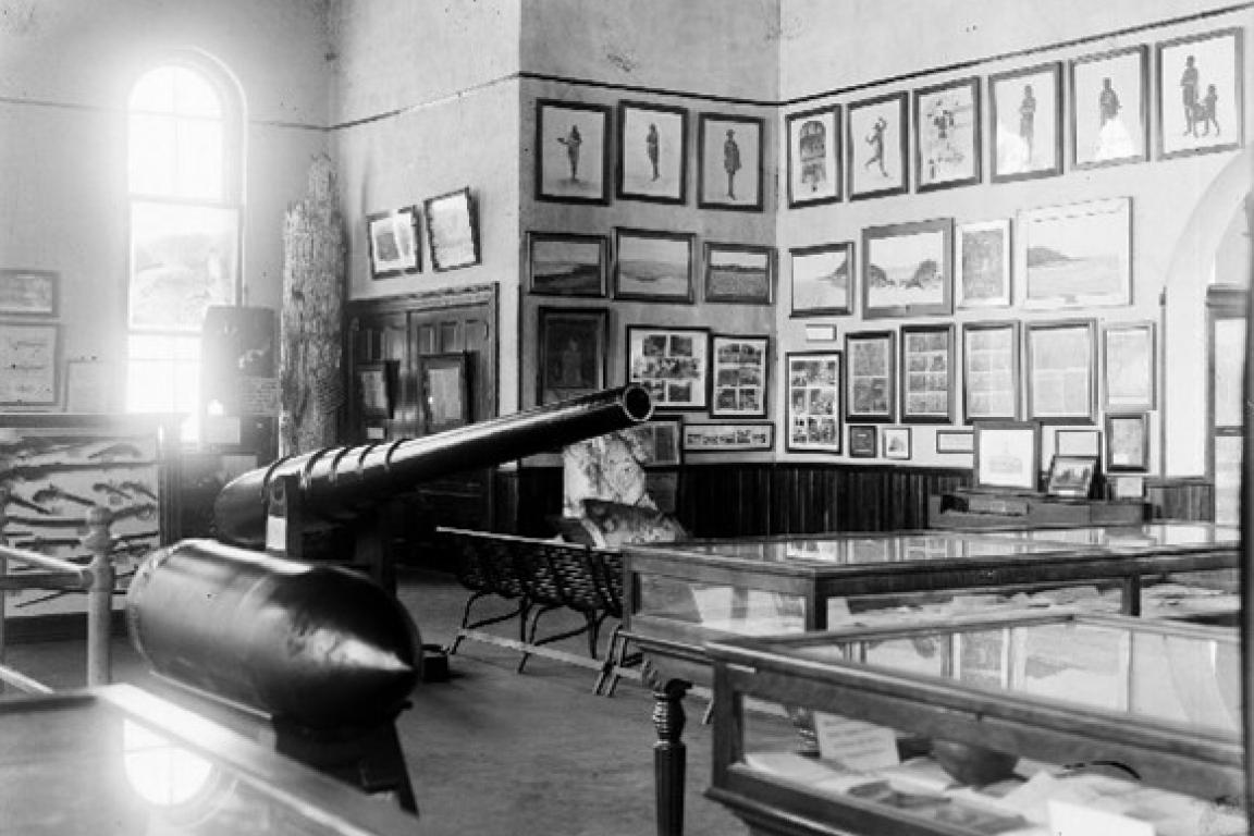 Black-and-white image of a historic museum gallery featuring a large cannon, glass display cases, and walls lined with framed images and documents.