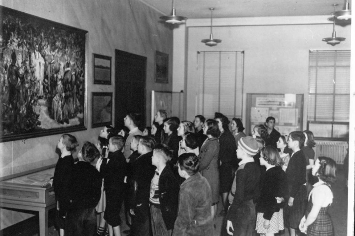 Black-and-white photograph of a group of schoolchildren gathered in a museum gallery, viewing a large painting on the wall.