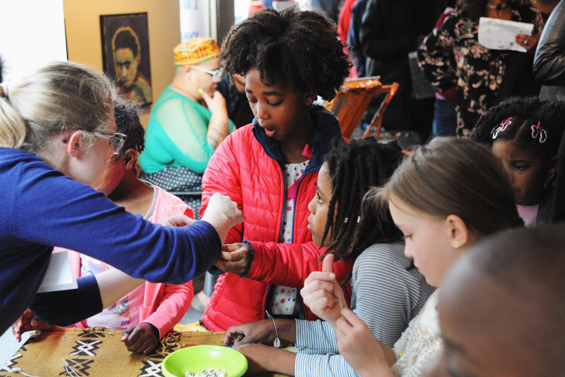 A woman assists a group of children gathered around a table for a hands-on museum activity involving string and small shells.