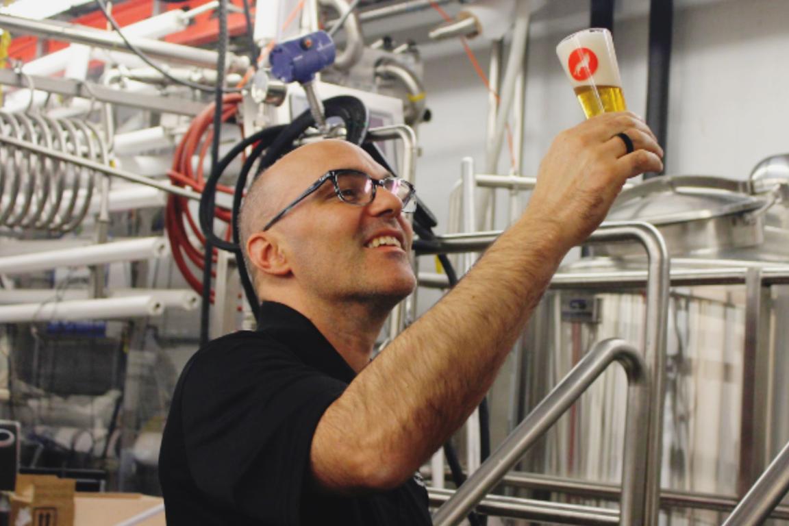 A smiling, bald man wearing glasses holds up a small glass of light-colored beer with a white head, examining it against the background of a commercial brewery with stainless steel tanks and piping.