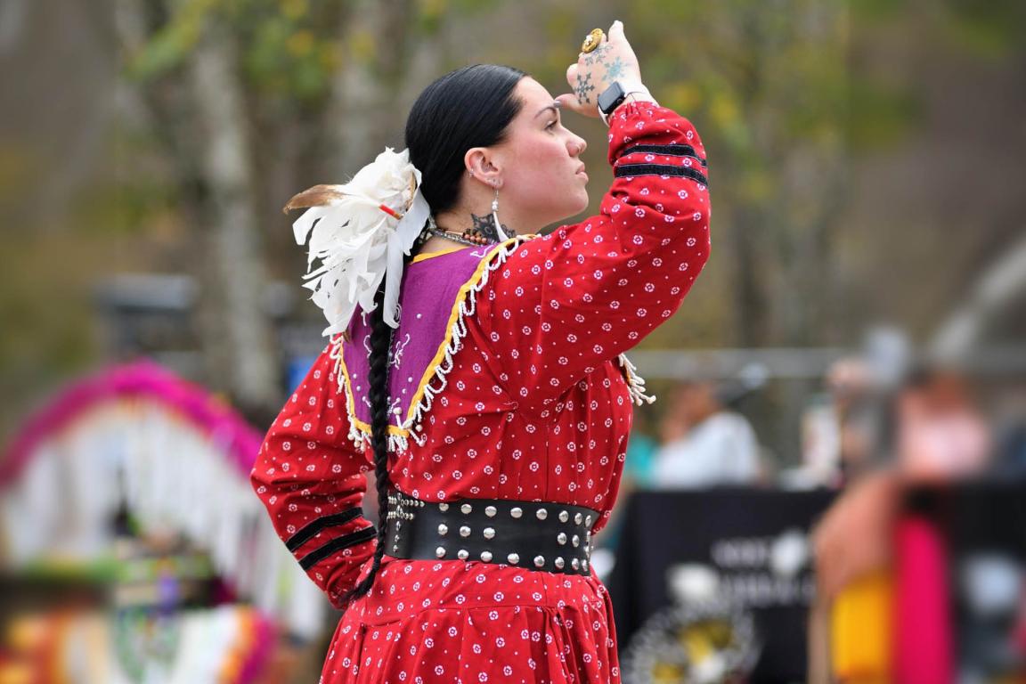Side profile of an Indigenous woman in a patterned red dress, purple beaded capelet, and wide studded belt. She is looking to the right with her hand raised to her brow, her dark hair styled in a braid with white feathers.