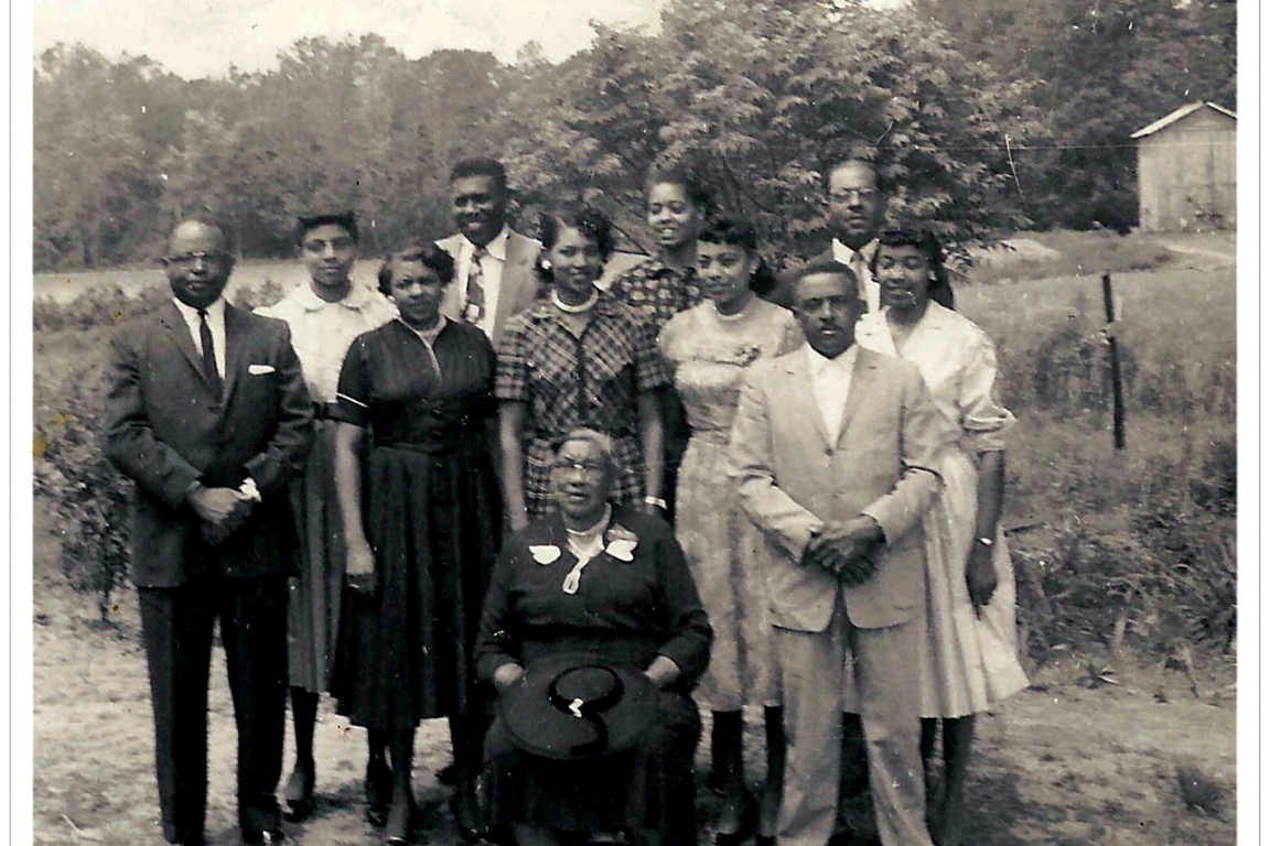 A black and white photo with a family standing together. An older woman sits in front. They are near a field with a line of trees in the back. 