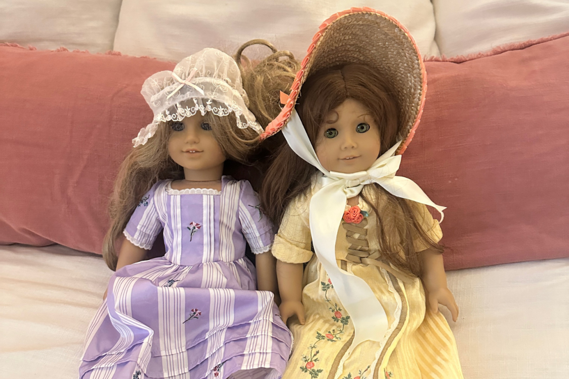 Image of two dolls side by side against a pillow. The left doll has a purple striped dress and a white bonnet. The right doll has a wide brimmed hat and a yellow dress. 