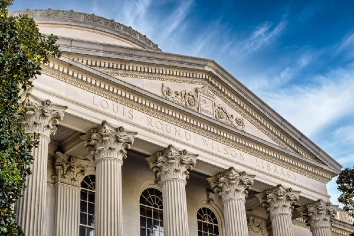 Facade of an old building with Greek columns in the front. The sky is blue and peppered with clouds. 