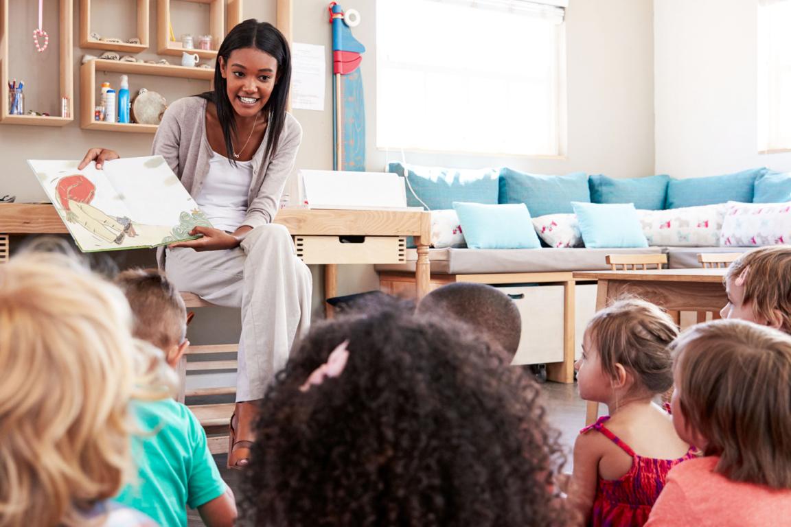 A woman with brown skin and long hair sits at the front of a room holding an open book. The back of children's heads can be seen in a crowd sitting on the floor. 