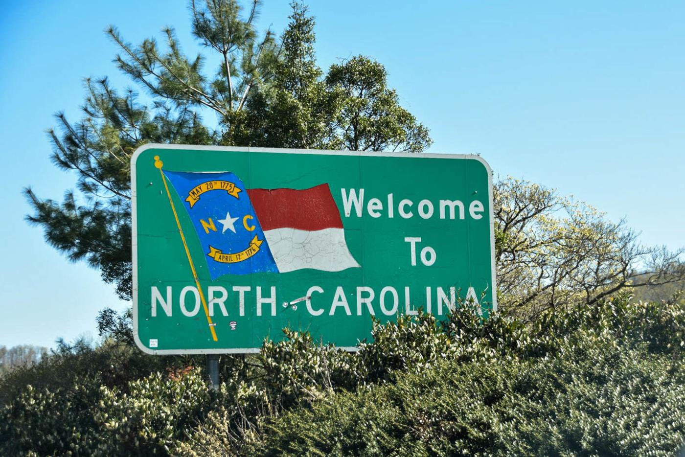 A green sign with the NC flag that says Welcome to North Carolina. The sign is nestled in green trees with a blue sky background. 