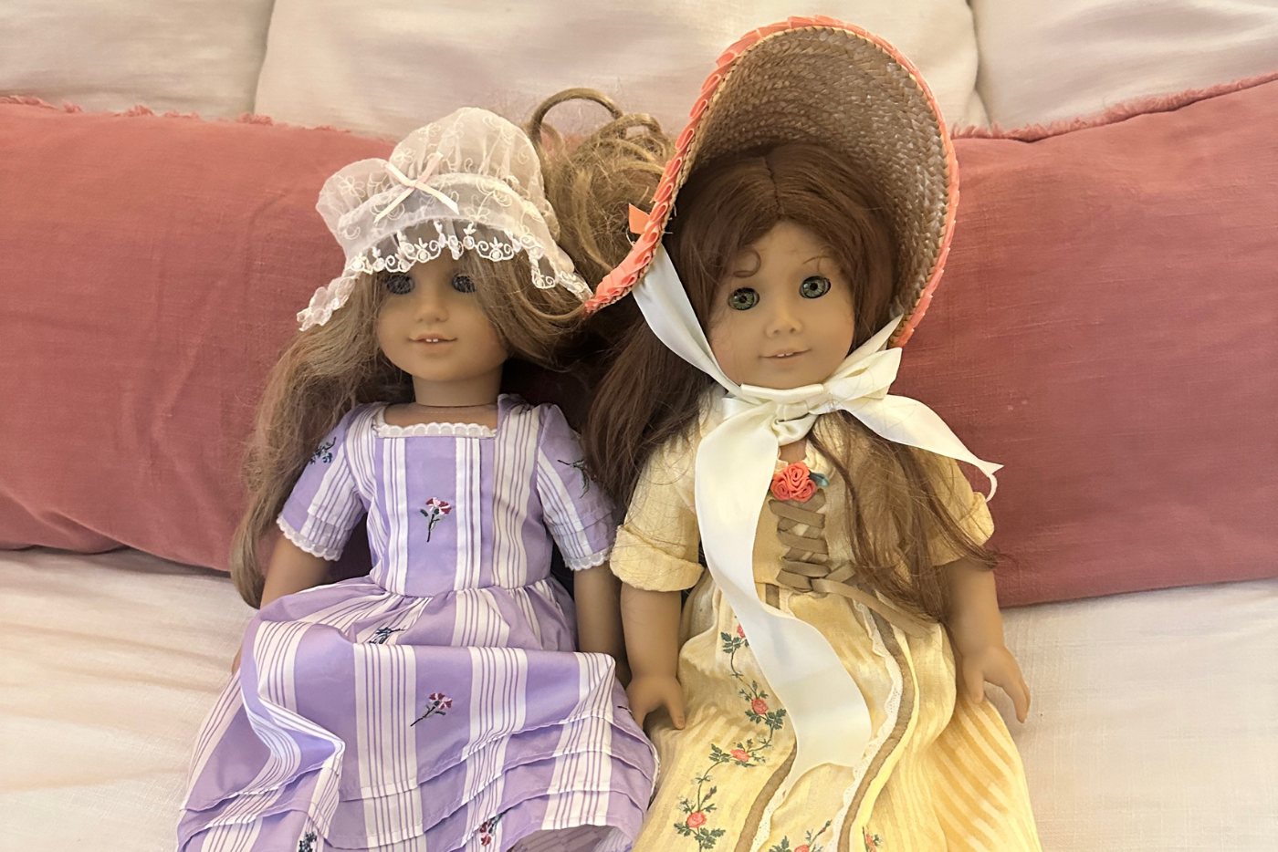 Twodolls sitting on a bed with pink and white pillows, one wearing a lavender dress and lace bonnet and the other wearing a yellow dress with a wide-brim straw hat.