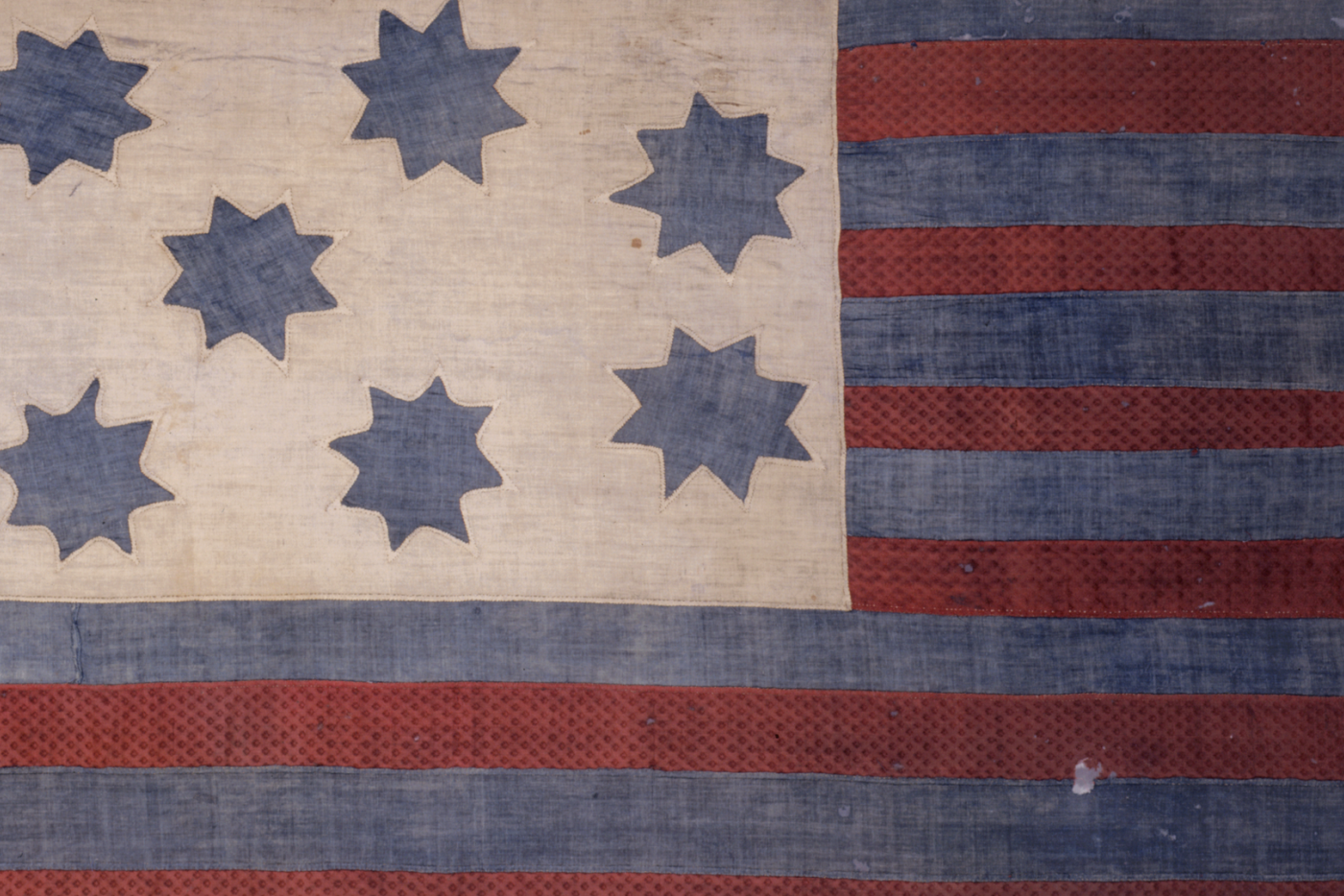 A close-up of a weathered, antique American Revolutionary War-era flag. It features a cream-colored canton on the left with seven blue, eight-pointed stars, and a field of alternating red and blue horizontal stripes on the right. The fabric is heavily textured and frayed, showing significant age.
