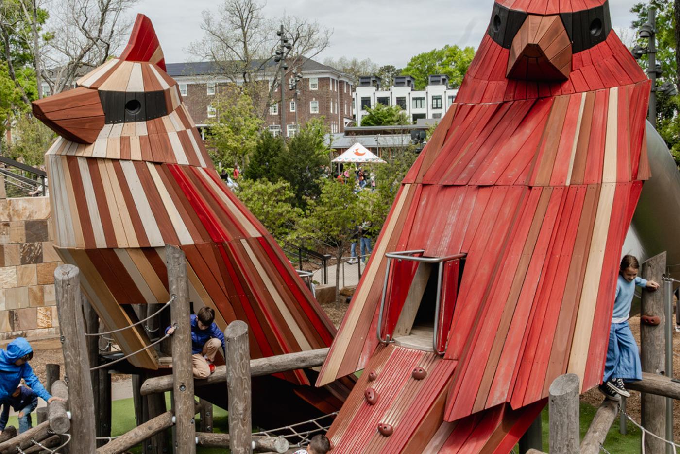 Two large, stylized wooden cardinal sculptures serve as the centerpiece of a multi-level outdoor playground where children are climbing and playing. The cardinals, one predominantly red and the other natural wood-toned, feature built-in slides and climbing handholds within a structure of logs and ropes.