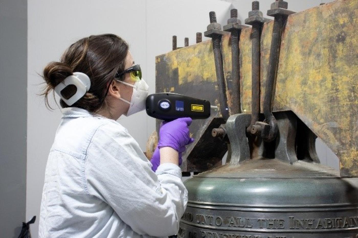 Museum conservator uses a handheld laser to remove corrosion from the Liberty Bell replica's metal frame during restoration.