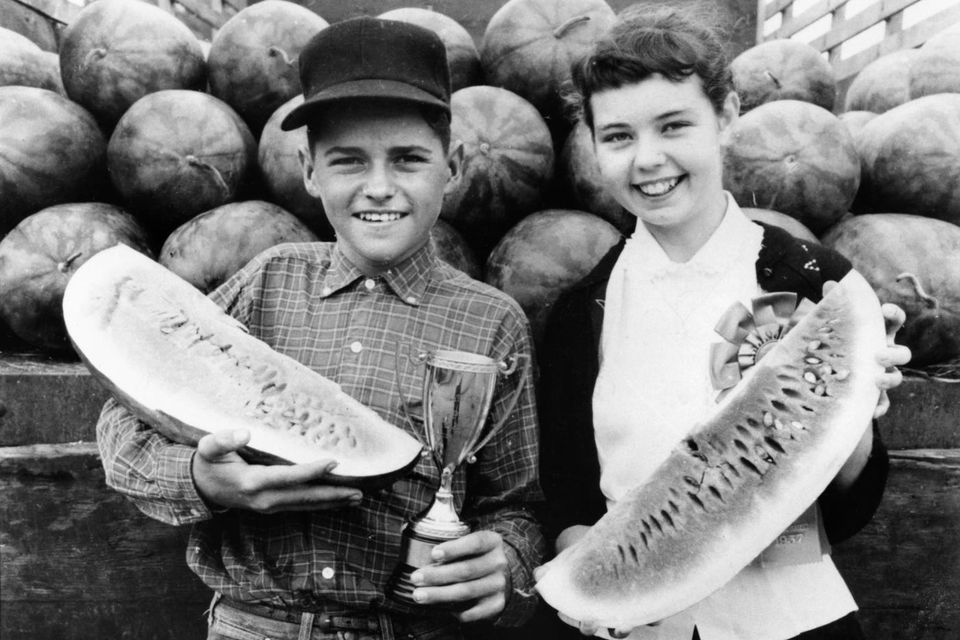 Two smiling children in a black and white photo. They are each holding a long piece of watermelon and stand in front of a pile of watermelons.
