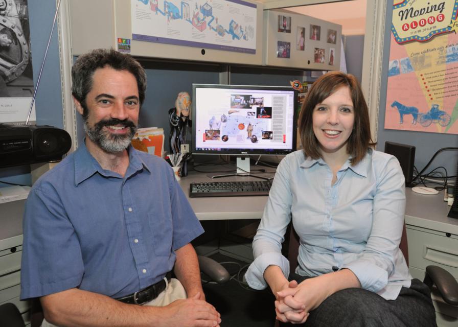 Two museum staff members seated in an office workspace with exhibit materials and a computer in the background.