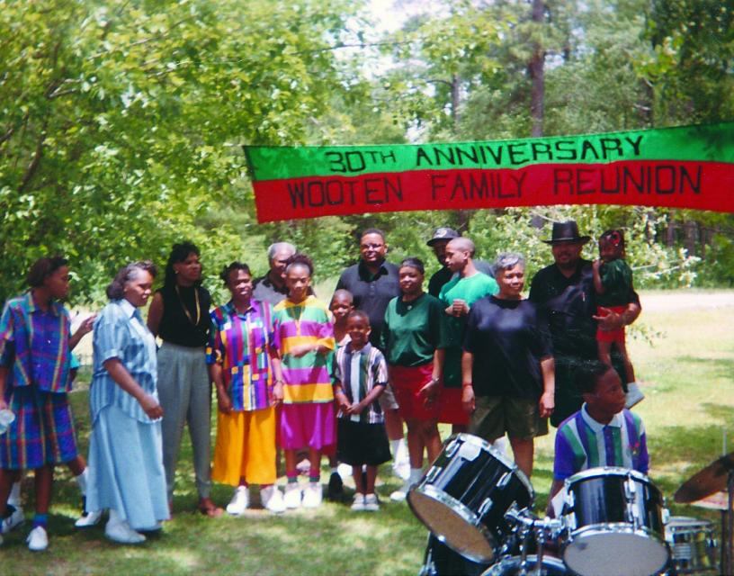 Photo of a family in a green field with a line of trees behind them. A banner reads "30th Anniversary Wooten Family Reunion"