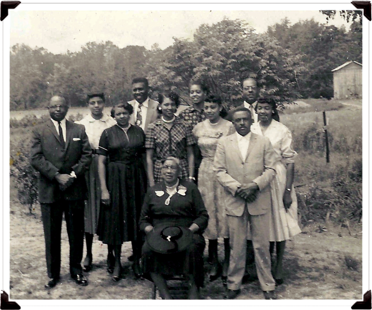 A black and white photo with a family standing together. An older woman sits in front. They are near a field with a line of trees in the back. 