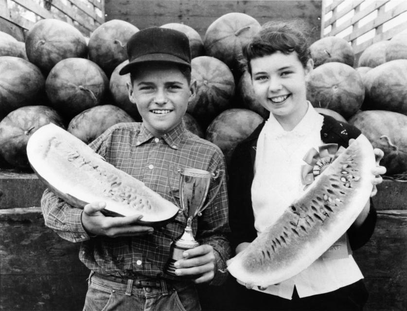 Two smiling children in a black and white photo. They are each holding a long piece of watermelon and stand in front of a pile of watermelons.
