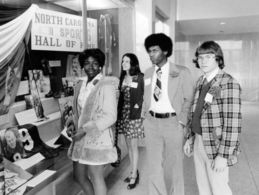 Four teens stand in front of a sports hall of fame display. Two look toward the camera and the other two look to the left.
