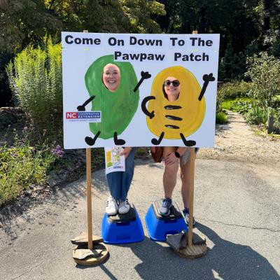Two people smiling with their faces in a cutout sign shaped like cartoon pawpaws at a Pawpaw Patch display.