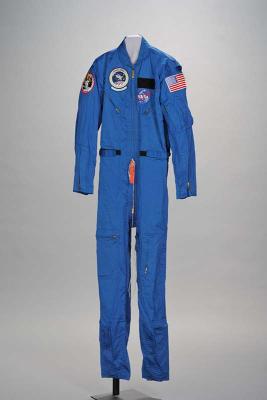A blue NASA flight suit displayed on a stand, featuring mission patches and an American flag on the sleeve.
