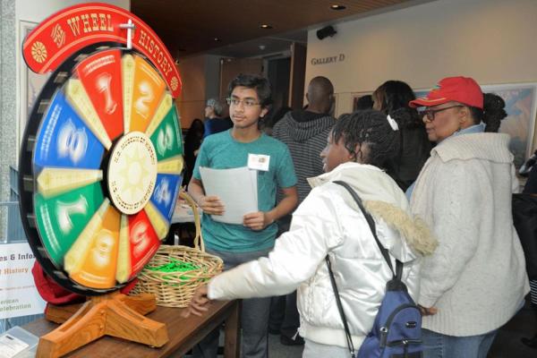 A group of museum visitors gathered around a colorful, numbered "History Wheel." A young person in a teal shirt looks on as another reaches toward a basket of prizes on the table.