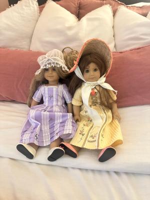 Two American Girl dolls sitting on a bed with pink and white pillows, one wearing a lavender dress and lace bonnet and the other wearing a yellow dress with a wide-brim straw hat.