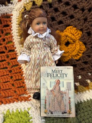 American Girl Felicity doll in a floral colonial dress standing beside a miniature copy of Meet Felicity, placed on a crocheted blanket.