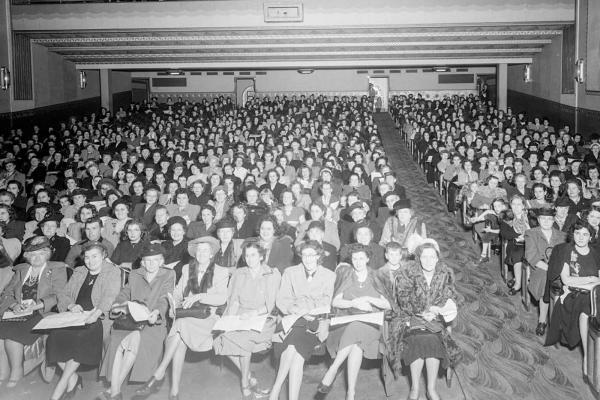 A black and white photograph from a high angle looking down at a packed audience of women wearing hats and coats in a classic mid-century theater. The women are all seated in long rows of chairs and appear to be waiting for an event to begin.