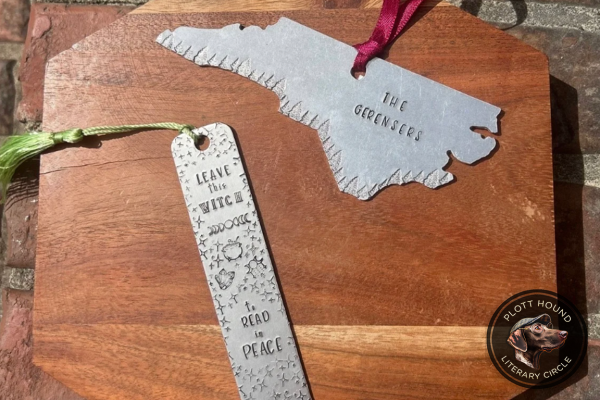 Two hand-stamped metal bookmarks—one shaped like North Carolina and another rectangular with a witch-themed inscription—resting on a wooden board.