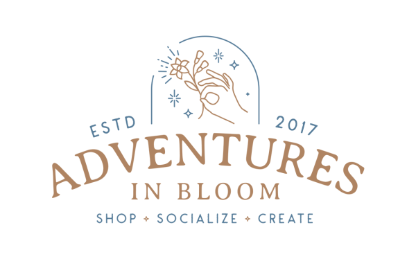 Logo for "Adventures in Bloom" featuring a hand holding a flower within a blue arch. Text includes "Estd 2017" and the tagline "Shop + Socialize + Create."
