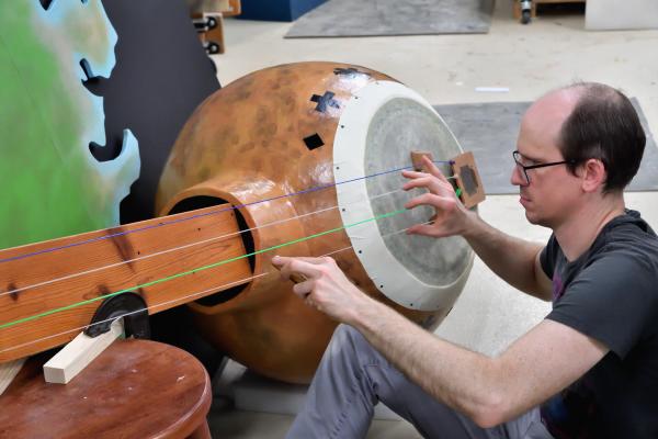Man adjusts strings on a large, handmade banjo mounted on a North Carolina-shaped backdrop in a workshop.