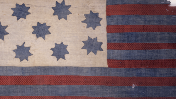 A close-up of a weathered, antique American Revolutionary War-era flag. It features a cream-colored canton on the left with seven blue, eight-pointed stars, and a field of alternating red and blue horizontal stripes on the right. The fabric is heavily textured and frayed, showing significant age.