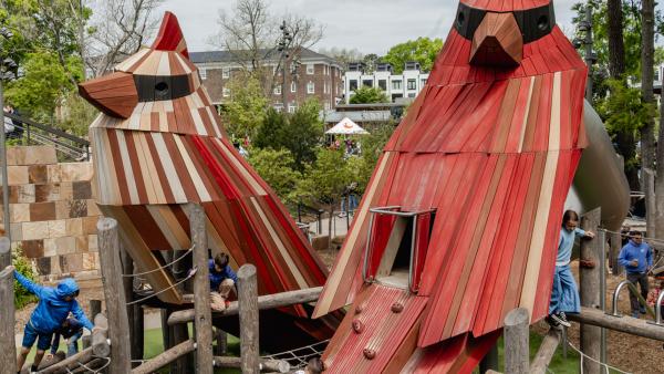 Two large, stylized wooden cardinal sculptures serve as the centerpiece of a multi-level outdoor playground where children are climbing and playing. The cardinals, one predominantly red and the other natural wood-toned, feature built-in slides and climbing handholds within a structure of logs and ropes.