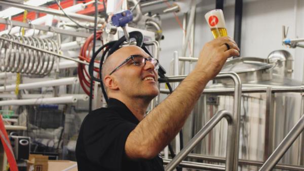 A smiling, bald man wearing glasses holds up a small glass of light-colored beer with a white head, examining it against the background of a commercial brewery with stainless steel tanks and piping.