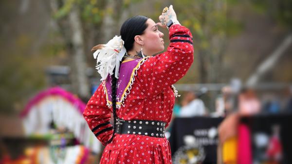 Side profile of an Indigenous woman in a patterned red dress, purple beaded capelet, and wide studded belt. She is looking to the right with her hand raised to her brow, her dark hair styled in a braid with white feathers.
