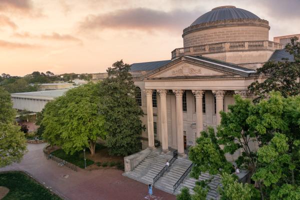 An elevated, wide shot of the Louis Round Wilson Library building during a warm sunset. The classical stone building features a prominent grand dome, a columned portico with wide steps, and is surrounded by lush green trees and a brick plaza.