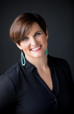 A brunette woman with short hair and turquoise earrings smiles at the camera. She wears a black shirt and sits in front of a dark gray background. 