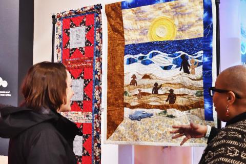 Two people discuss a colorful quilted artwork in a gallery.  