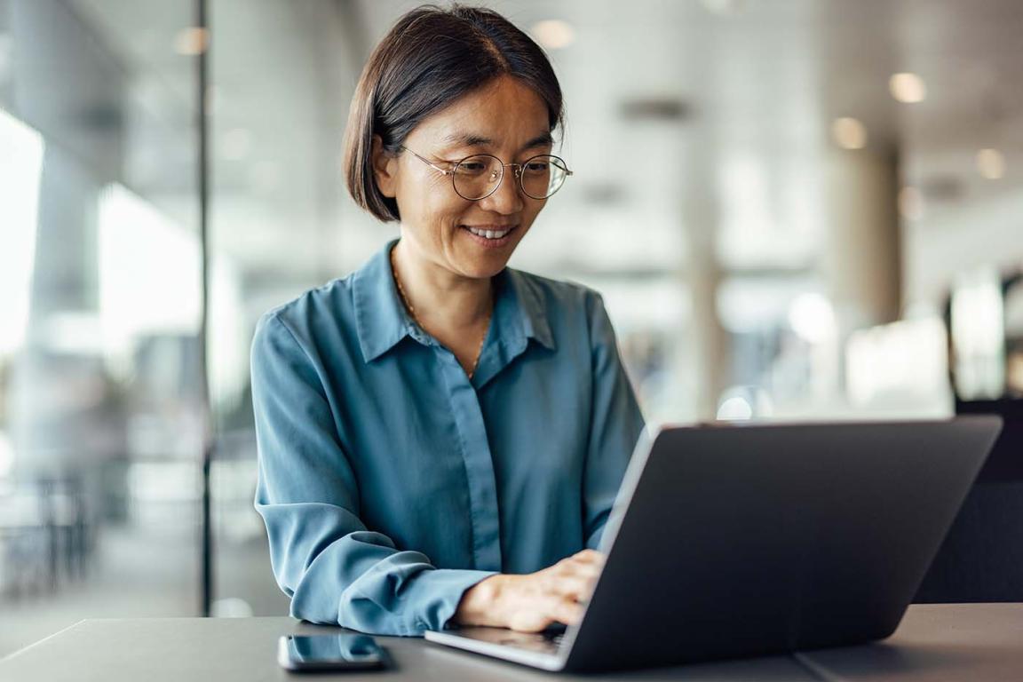 A middle aged Asian woman smiles and types on a laptop in a bright, airy office.