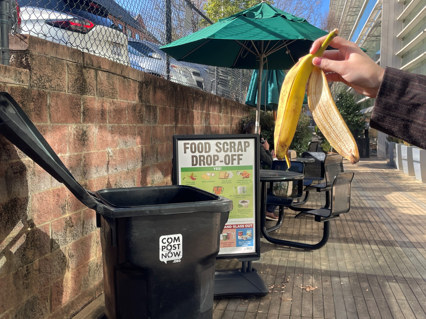 A compost bin is in front of a Food Scrap Drop-Off sign and a hand holding a banana peel is in view