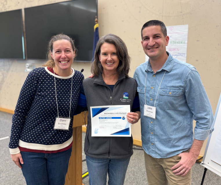 Three people standing for a photo with the person in the middle holding a certificate