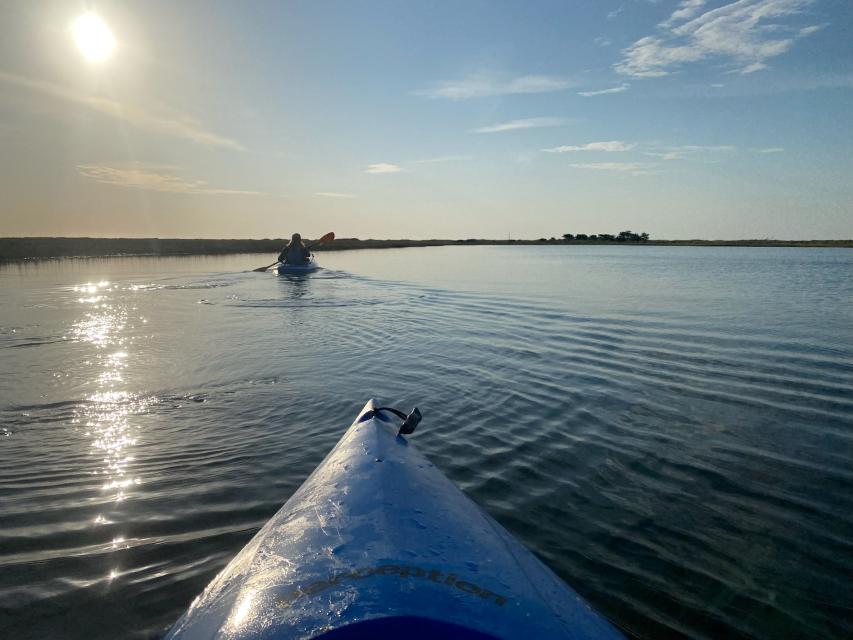 Someone kayaking on flat water into the sun with marsh surrounding them.