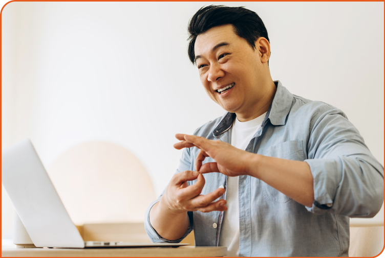A middle-aged Asian man smiles using sign language to communicate via a videoconference on his laptop.