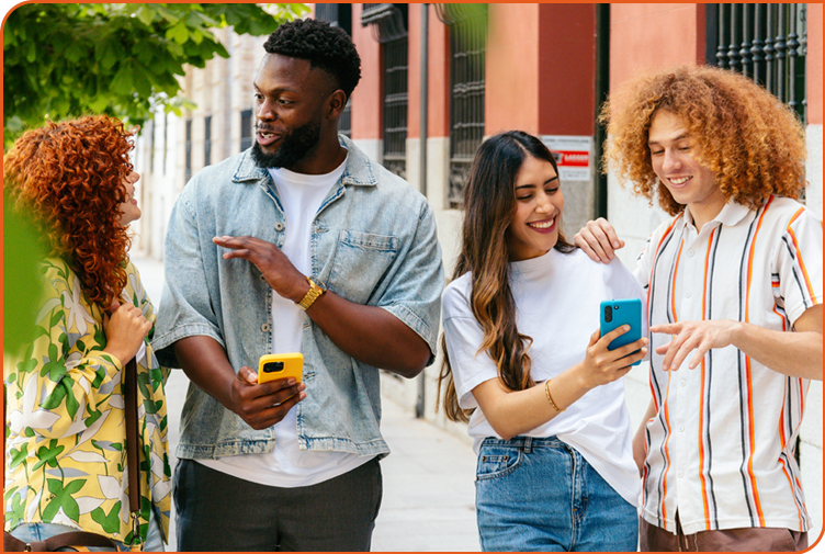 A diverse group of 4 young adults stands on a city sidewalk chatting about social media content on the smartphones they are holding.