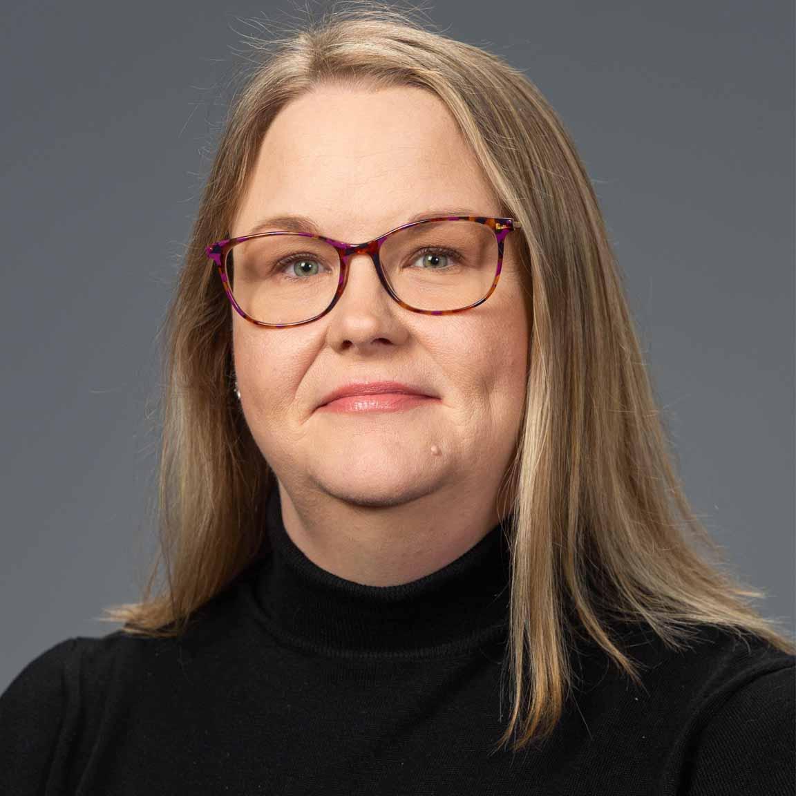 A white lady in her mid 40's with straight, dirty blonde hair and dimples. Wears a black turtleneck and eyeglasses, smiles.