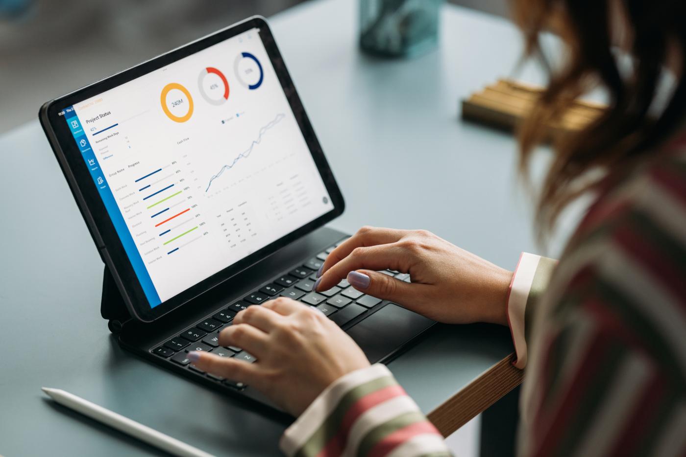 Woman working on a tablet displaying colorful graphs and data with a small keyboard in a business environment.