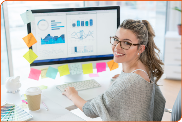 A young female marketing and communications strategist turns to smile at the camera; over her shoulder is a computer monitor displaying data trends.